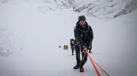Tim Howell Tim Howell is climbing up a mountain holding on to rope. He is wearing weather protective gear. There is a team of people behind him.