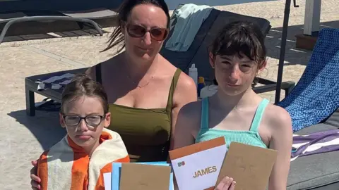 Melanie Luton A group of three people - a mother and her young son and daughter - stand outdoors by a poolside area, holding several cards and envelopes—one prominently labelled “JAMES”—while sun loungers and a paved patio are visible in the background.