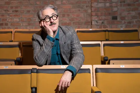 PA Media Alan Cumming sits in a theatre. He has short grey hair and wears glasses and a suit