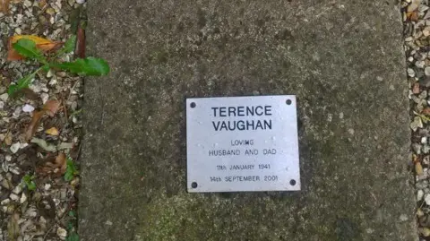 Carol Vaughan Close up image of a plain slab with a small metal plate, which has a name and four lines of writing underneath