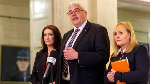 PA Media Three adults stand indoors at a press briefing, with a man with grey hair in a dark suit and striped tie speaking at a microphone bearing an official NI Assembly crest. Two women stand on either side - one with long brown hair in a dark blazer and another blonde haired woman holding an orange folder, all stood against a backdrop of light-coloured columns and framed artwork.

