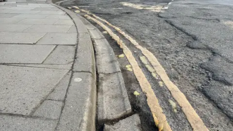 A Wiltshire road with an uneven surface with the top layer of tarmac broken up. The gutter running next to the pavement is broken and the yellow lines are patchy