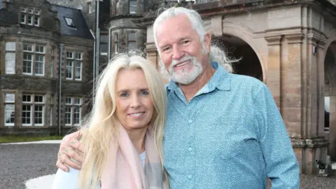 Suzanne and Lance Holmquist standing outside Carbisdale Castle. Suzanne has long blonde hair and is wearing a pink scarf over a white top. Kance is tall man with white hair and beard.