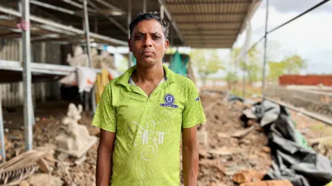 Shardul Kadam Dilip Mhatre stands in a green t-shirt, behind him is a blurred backdrop of his damaged workshop