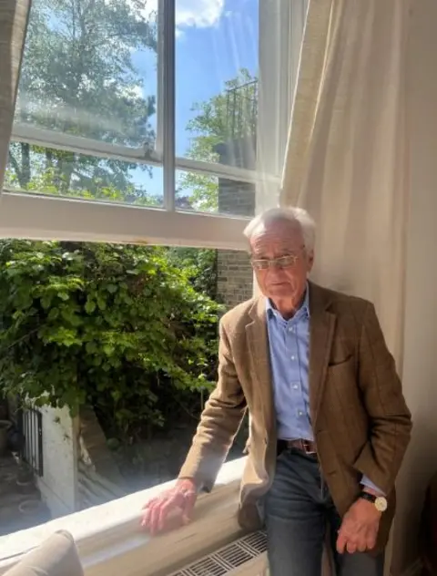 LDRS An older person standing indoors by a large window, with thick vegetation visible outside filling the neighbouring garden.