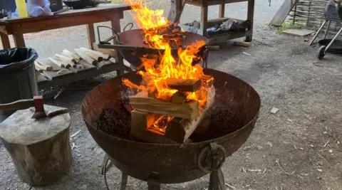 Fire is visible in a bowl which is above a larger bowl that is outside. A bench has objects on in the background and a bin is nearby.