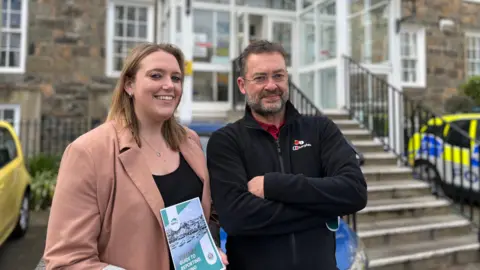 On the left hand side Poppy Murray, wearing a pink blazer and black top smiling holding the green guide to reporting spiking. She is stood next to Simon Allum who is wearing a black fleece jumper with his arms crossed. He is wearing glasses. In the background a blurred police vehicle. They are stood outside the steps the the Guernsey Police Station. 