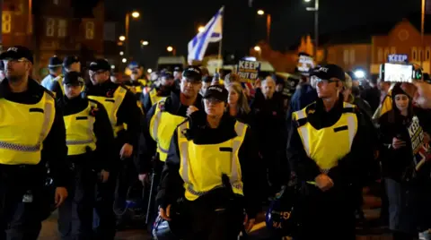 PA Media A group of officers are walking down a street outside Villa Park, most are male but in the middle is a white female officer wearing a black cap, all of them are wearing hi-vis vests.
