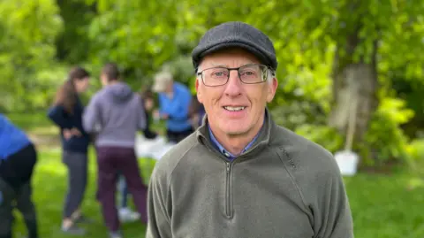 A man with a bonnet and glasses, wearing a dark green top, looks at the camera