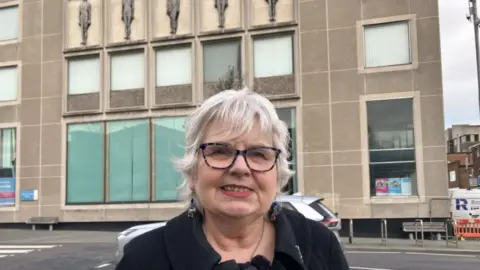 lara king short grey haired woman smiling, she's wearing glasses and has on a dark coat. She is standing next to a road in front of a large building with big windows. There are pieces of art on the side of the building of five male figures which are grey.