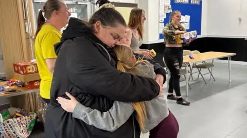 Simon Thake A woman with dark hair and a black coat gives a big bear hug to a younger girl in a grey top