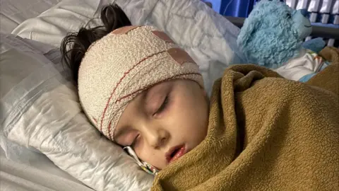 A little girl lies asleep on a hospital bed. Her head is bandaged and she is wrapped in a brown fleece blanket. Next to her is a blue teddy bear.