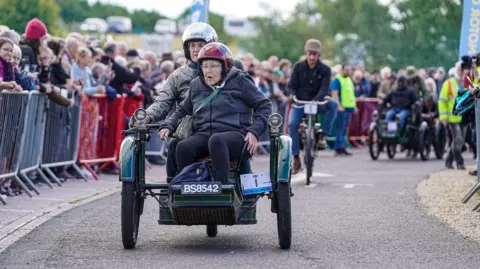 Peter Wileman A vintage three-wheeled vehicle being ridden by a woman wearing a crash helmet. Sitting in front of her as a passenger is an elderly woman, also wearing a crash helmet and glasses. They are travelling along a road lined with crowds of people standing behind a barrier.