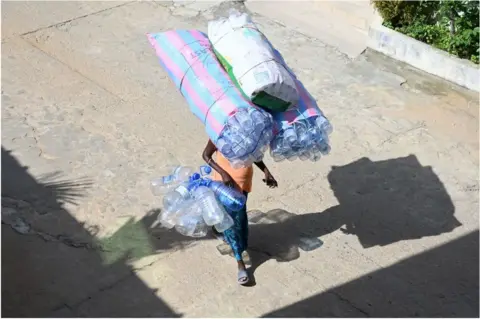ISSOUF SANOGO/ AFP A woman carrying plastic bottles on her head whilst walking down the street.