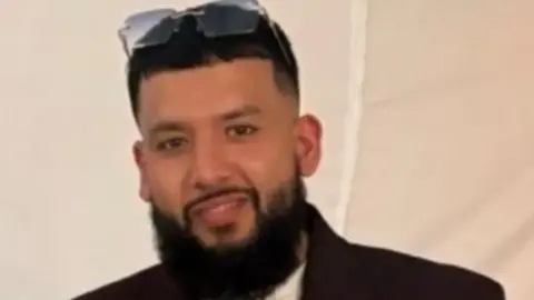 Ali is smiling at the camera and wearing dark clothing and has sunglasses on his head. He has a full beard.