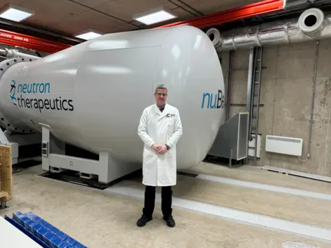 Professor Jason Parsons standing by the neutron accelerator, at the University of Birmingham. He is wearing a white lab coat and black trousers and shoes.