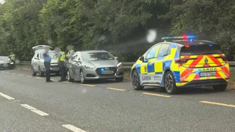 Garda officers and garda cars surround a small car on the roadside of the N2 near Carrickmacross. The officers are all wearing blue uniforms, but two are wearing yellow high-viz vests over their shirts. The boot of a hatchback vehicle behind them is open. 