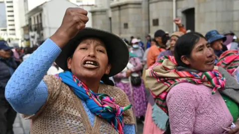 A woman shouts and shakes her fist while participating in a march calling for the resignation of Bolivian President Rodrigo Paz in La Paz, on 25 May, 2026. In the background, other protesters can be seen. Some are wearing colourful indigenous shawls.