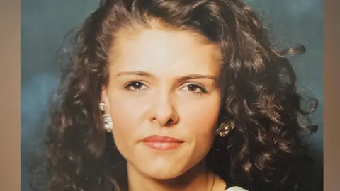 A close-up photo of a woman with brown curly hair , wearing jewellery and a black and white striped top.