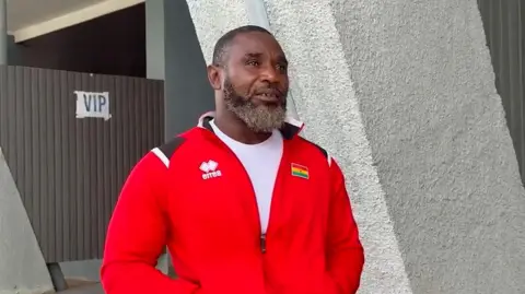 Head and body image of Raphael Botsyo Nkegbe. He is black with short dark hair a medium length grey beard. He is wearing a bright red zip top with a small Ghana flag and a white t-shirt. He has his hands in the zipper pocket.