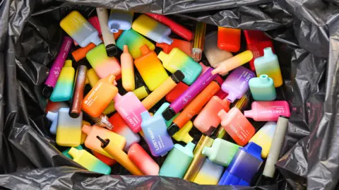 A large pile of multi-coloured vapes sitting in a black bin liner.