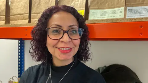 Zainab Aboharoon smiles at the camera. She is wearing burgundy glasses and a black jumper. 