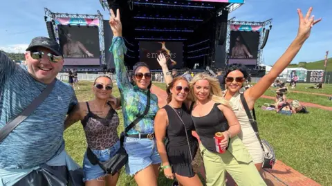 Six festival-goers - one man and five women - smile into the camera as they stand in the area near the large stage where musicians will perform.