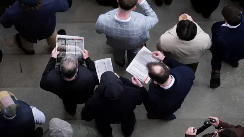 PA A picture taken from above of five men in the centre of the image, standing on the concrete steps in the grandstands at Cheltenham Racecourse. They are studying the form, which looks like a newspaper, where all the horses' previous race statistics are detailed.