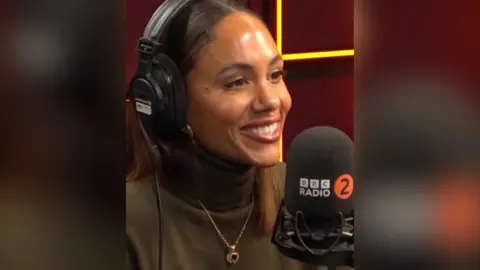 Alex Scott has long brown hair and wears a brown turtle neck. She has headphones on and is in a radio studio