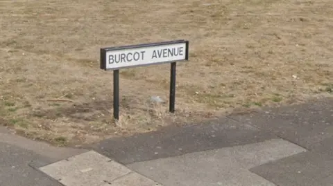 Google Dry grassy area with a sign which reads Burcot Avenue above. There is pavement to the front of the image