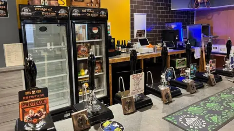 Andy Evans A bar, with beer mats placed along the surface and fridges and counters behind. The walls are a mixture of dark yellow and grey tiles in a brick effect. There are seven handles to pull pints and tills in the background too. 