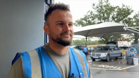 Image shows Klodian looking off to the side of the image. He is wearing a blue high-vis and a grey top underneath. He has dark brown hair. Behind him is the car washing area. There is a white canopy and green trees. Underneath the canopy, are two cars getting hoovered and polished. There are workers in the background in blue high vis.