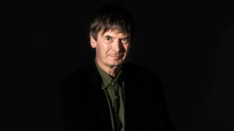 A man wearing a dark jacket and a green shirt looks at the camera