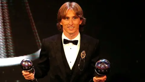 Reuters Luke Modric is seen in a tuxedo, holding two awards - one in each hand - in the middle of an awards ceremony