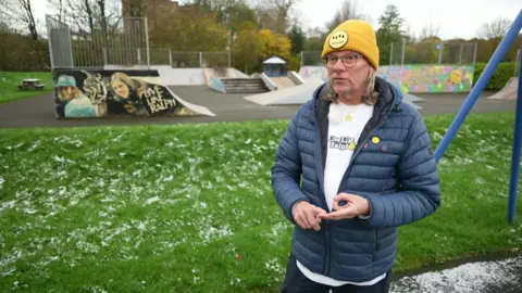 Neil is stood at skatepark and is wearing a blue winter jacket and white t-shirt with the text Live Like Ralph