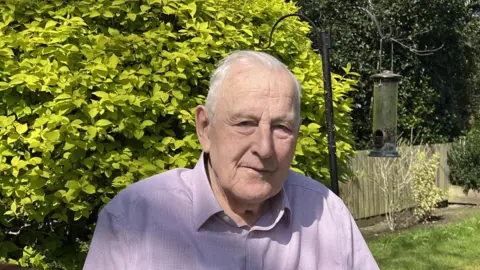 Peter Walker from Pewsey looks at the camera from his garden, a bird feeder and green hedge in the background. He wears a shirt.
