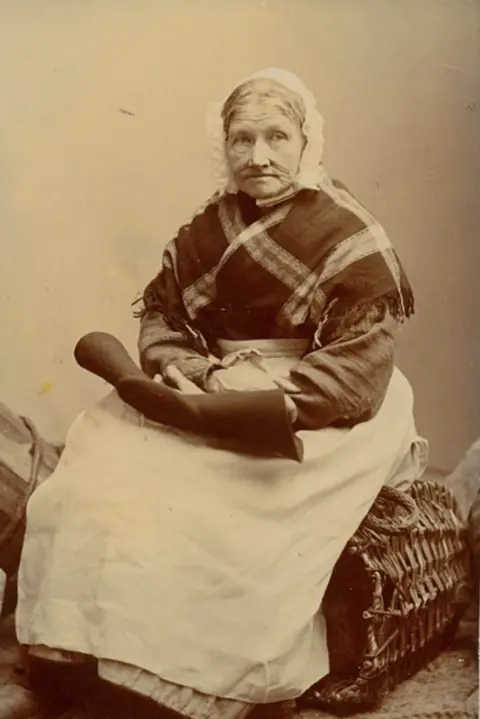 Amlwch Industrial Heritage Trust A sepia photograph showing an old lady sitting on a wicker basket with her gauntlet on her knee. She is wearing a white apron and a lace cap with a shawl around her shoulders