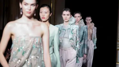 Getty Images Models walking on a catwalk, all with dark slicked-back hair, wearing outfits in silky fabrics in mint green and baby pink, some accessorised with bows on their shoulders 