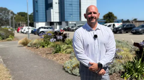 Luke has a bald head and wears a white shirt and grey trousers with his arms crossed across his chest. He stands in the college grounds with parked cars and a tall glass fronted building partially visible behind him.