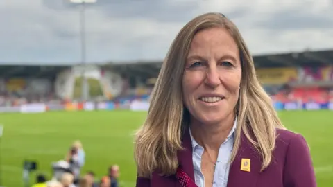 Sarah Massey Sarah Massey wearing a burgundy blazer and a light blue blouse, with her blonde hair over he shoulders and smiling lightly at the camera. 
She is standing in front of a rugby stadium, with lots of green grass, with the background out of focus. 
