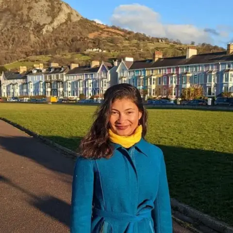 Leena Farhat A woman with long dark hair wearing a yellow scarf and blue coat, stood on a path with a patch of grass and row of colourful houses bbehind her. Behind the houses is a mountain.