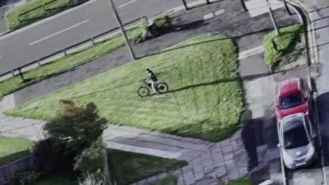 Drone footage of a man riding a stolen bike in north Manchester. The image shows a man riding on grass, close to a road and parked cars.