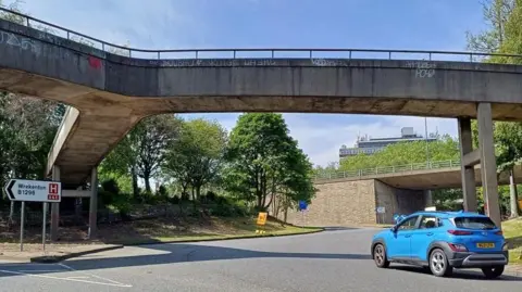 A blue car travels around the Five Bridges roundabout. The roundabout's concrete bridges stand above it, and splits off into multiple directions. The Gateshead flyover can be seen in the background.