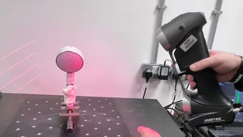A handheld 3D scanner is used to scan a shower head mounted on a stand on a laboratory bench, with red laser points visible on the object and surface. There is a moulded hand shape made of plasticine around the handle. 