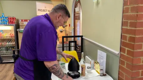 Alex Pope/BBC Stan working in the cafe, leaning over and cleaning a table, with items on it. He is side-on to the camera wearing an apron, purple top. He has tattoos on his right forearm. 