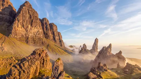 John Dewar A dramatic mountain landscape with towering rock formations, steep cliffs, and rolling grassy slopes, on a misty but sunny morning.