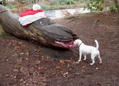 George Burgess Large wooden "snake shaped" log with eyes painted on and red glitter painted to look like lipstick wearing a large Santa hat with a small white dog looking into the "mouth".