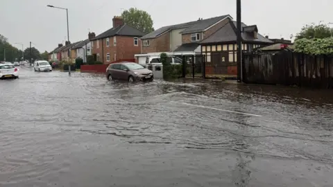 Asim Mahmood A car is parked on the side of a road which has been flooded. Large volumes of water are on the road. The water has gone on to the pavement and is near a row of houses. 