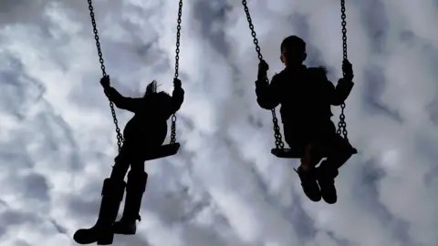 PA Media Two silhouettes of children swinging on swings. The sky is in the background.