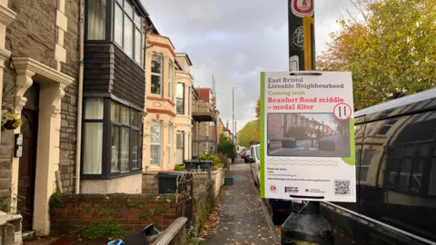 A poster attached to a lamppost on a residential street reads “East Bristol Liveable Neighbourhood Coming Soon – Beauford Road middle -Modal Filter,” printed in black, green and red text on a white background.